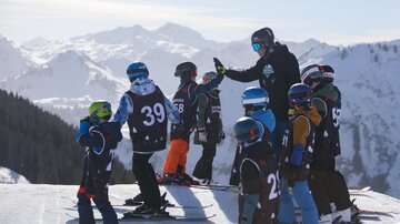 A group of children is standing on skis and looking at a teacher who is giving them instructions. In the background, there are snow-covered mountains and a clear sky.