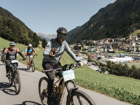 A group of cyclists on a picturesque path in the mountains. In the background, a small village surrounded by green meadows and mountains can be seen.