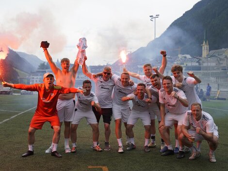 A group of soccer players is celebrating on the field. They are cheerful and holding torches in their hands, while a picturesque mountain landscape can be seen in the background.