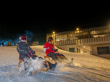 Two people are riding a sled on a lit snow slope at night. In the background, a building can be seen.