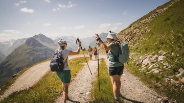 A group of hikers is on a hiking trail in the mountains. The landscape is green with mountains in the background and a clear sky.