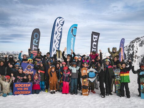 A group of children and adults stands happily in the snow. They are wearing ski equipment and are celebrating with flags in the background.