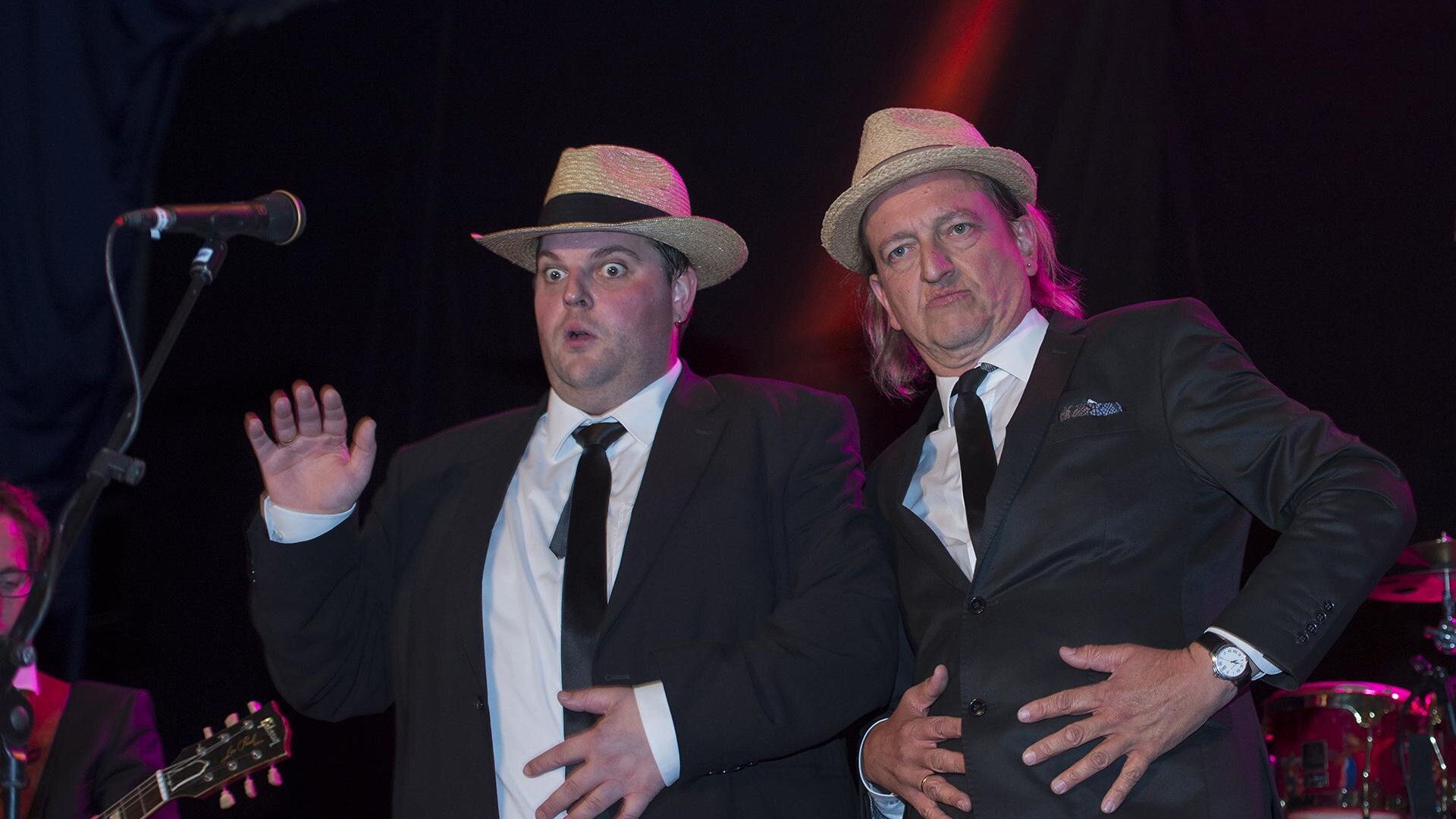 Two men in suits and hats are posing amusingly on stage. In the background, musicians with instruments can be seen.