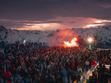 A large crowd gathers in the mountains at sunset. Festive atmosphere with colorful lights and smoke torches.
