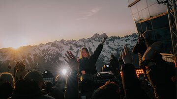A DJane plays music on a stage with a breathtaking view of snow-capped mountains. The crowd dances and celebrates at sunset.