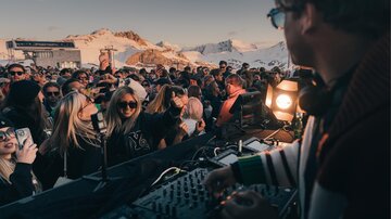 A lively outdoor party with many people and a DJ. Snow-covered mountains can be seen in the background.