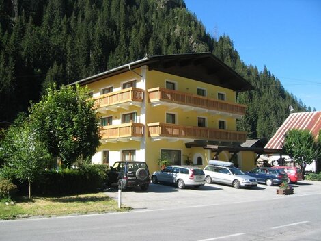 A bright building with several balconies, surrounded by trees and mountains. Several cars are parked in the parking lot.