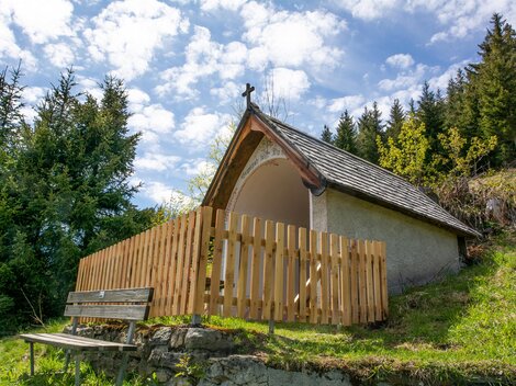 A small chapel with a wooden fence stands in a wooded environment. The sky is partly cloudy and the landscape appears calm and peaceful. | © Bernhard Gruber – Photography