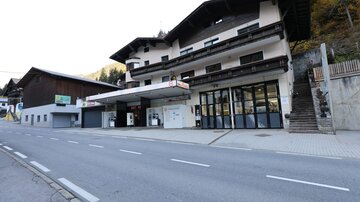 A building with a gas station and sales rooms on a quiet street. In the background, additional traditional alpine buildings are visible.