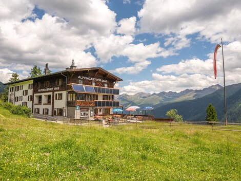 A picturesque mountain cabin surrounded by green meadows and mountains. The sky is partly cloudy and the atmosphere is inviting.