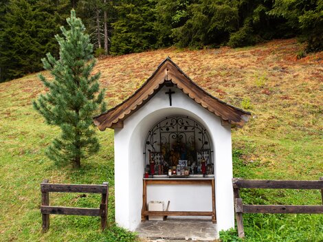 A small wooden shrine stands in a green field, surrounded by trees. The shrine has a red roof and is decorated with a cross and small statues. | © Bernhard Gruber – Photography