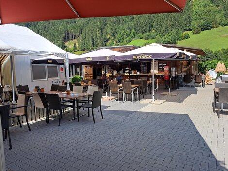 A cozy outdoor area of a restaurant with tables and chairs. In the background, there is the restaurant with umbrellas and a green landscape.