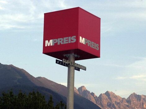 A red advertisement sign with the inscription "MPREIS" stands in front of a mountain horizon. The sign points to a road with the addition "Supermarket".
