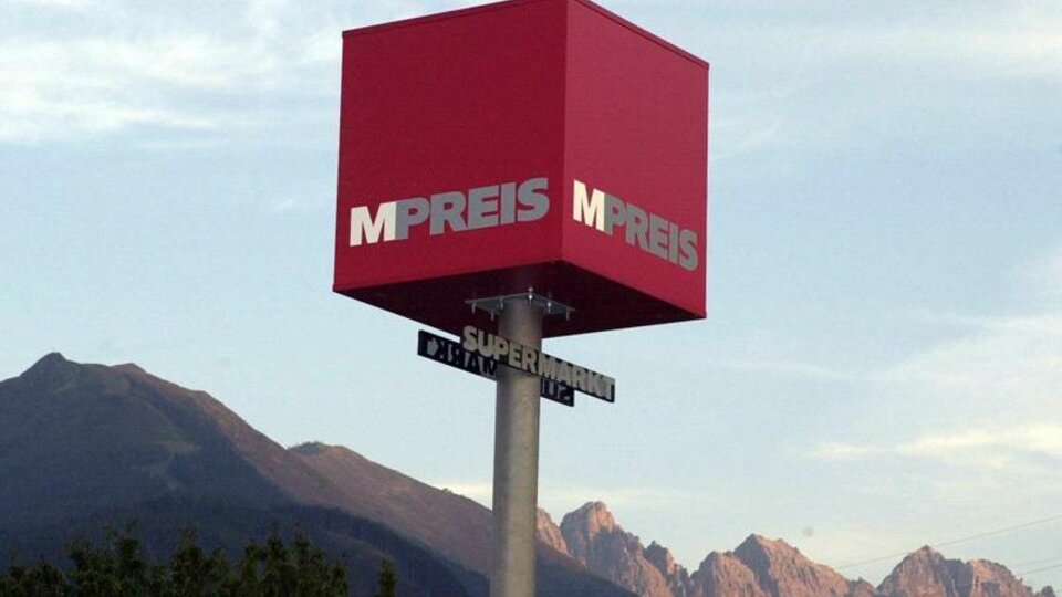 A red advertisement sign with the inscription "MPREIS" stands in front of a mountain horizon. The sign points to a road with the addition "Supermarket".