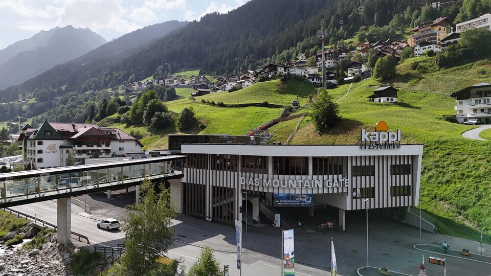 Ein modernes Gebäude mit der Aufschrift "Kappl" in einer bergigen Landschaft. Im Hintergrund sind grüne Hügel und kleine Häuser zu sehen.