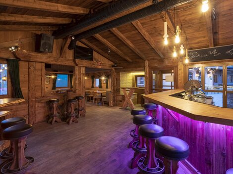 A cozy bar area with wooden interior and soft lighting. The bar has a modern, purple-lit surface and some seating.