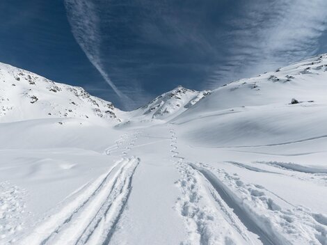 A snowy landscape with tracks in the snow and high mountains. The sky is blue with some clouds.