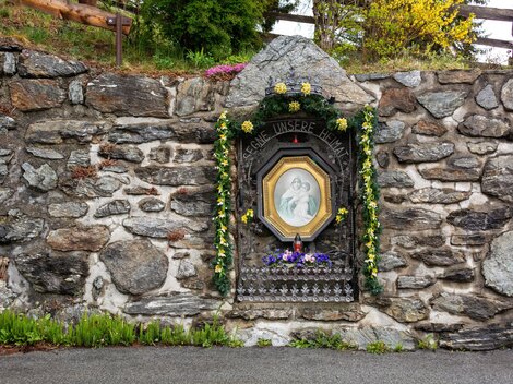 A stone wall with a decorated portrait in a niche. The image is adorned with flowers and depicts a religious scene. | © Bernhard Gruber – Photography