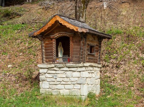 A small wooden chapel stands on a stone pedestal in nature. Next to the chapel, green meadows and foliage can be seen. | © Bernhard Gruber – Photography