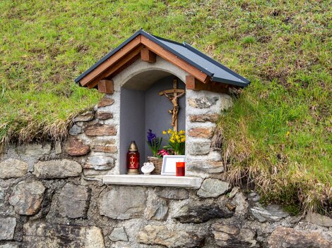 A small niche in a stone wall with a wooden roof. Inside are figures, candles, and flowers for devotion. | © Bernhard Gruber – Photography