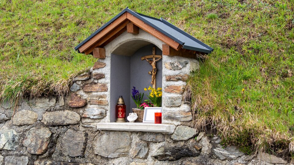 A small niche in a stone wall with a wooden roof. Inside are figures, candles, and flowers for devotion. | © Bernhard Gruber – Photography