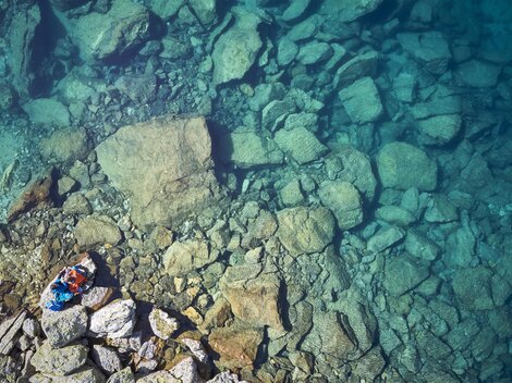 A clear blue lake with many large stones at the bottom. A person is lying relaxed on the shore.