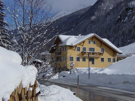 A charming yellow house in the snow with a picturesque mountain backdrop. The path is covered in fresh snow and the trees are decorated for winter.