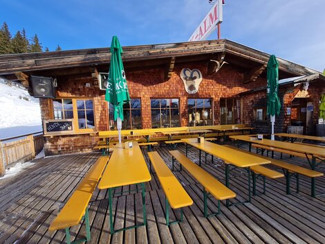 A cozy mountain cabin with wood cladding and large windows. Outside, there are long benches and tables with green umbrellas.