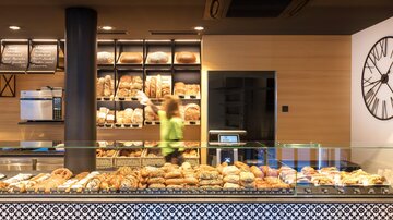 A modern bakery with a selection of fresh bread and pastries. In the background, an employee is seen working.
