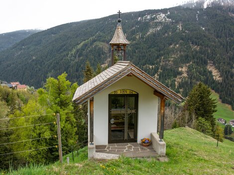 A small chapel stands on a hill, surrounded by trees and mountains. In the background, a picturesque mountain landscape can be seen. | © Bernhard Gruber – Photography