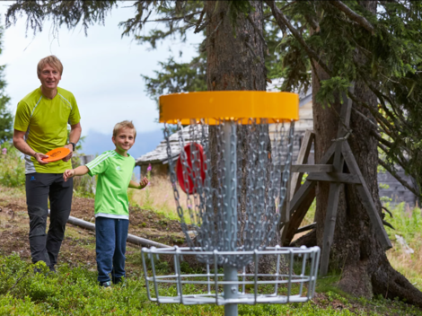 A man and a boy are standing in a forest and playing disc golf. In the background, the basket for the game can be seen.