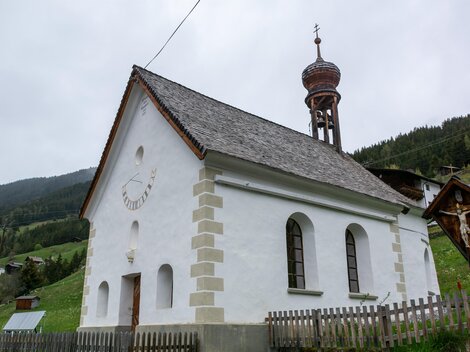 A small, white church with a tall tower stands in a green landscape. In the background, gentle hills and forests can be seen. | © Bernhard Gruber – Photography