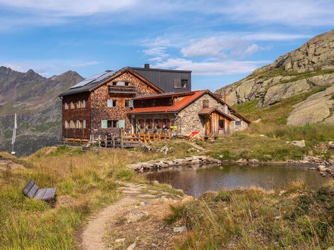 A picturesque mountain cabin in the midst of a green landscape. In the foreground, there is a small pond, and in the background, impressive mountains can be seen.