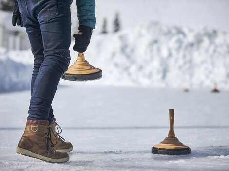 A person is standing on a frozen surface holding a wooden block in their hand. Snow-covered hills are visible in the background.