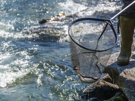 A fisherman stands on the shore of a body of water and holds a net in hand. The water flows sparkling in the background.