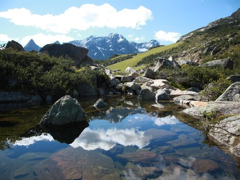 A picturesque mountain landscape with high peaks and a clear blue sky. In the foreground, the terrain is reflected in the calm water of a small pond.