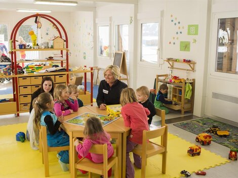 A bright kindergarten room with children sitting around a table and playing. A caregiver is present, while in the background, other children are occupied with toys.