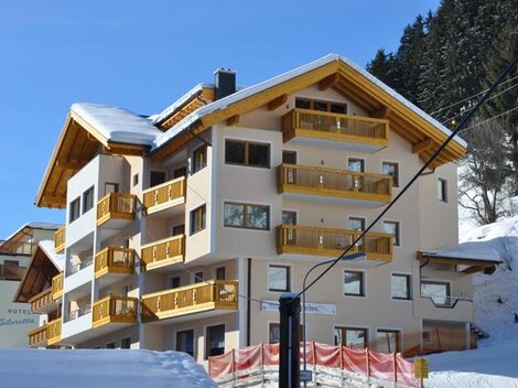 A modern hotel building with wooden balconies in a snowy environment. In the background, there are fir trees and a clear blue sky.
