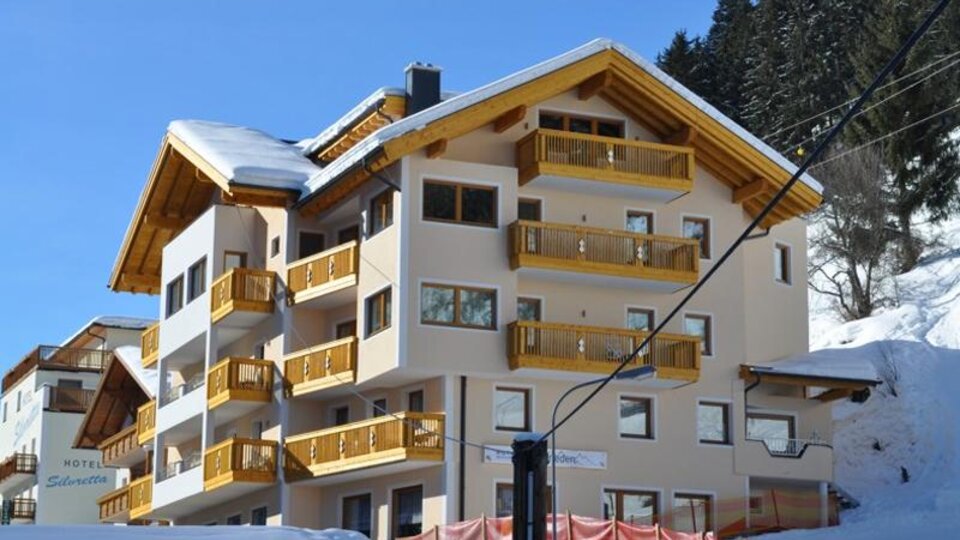 A modern hotel building with wooden balconies in a snowy environment. In the background, there are fir trees and a clear blue sky.