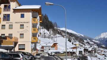 A hotel in a snowy mountain landscape, surrounded by snow-covered mountains and small houses. The road in front of the hotel is narrow and easily passable.