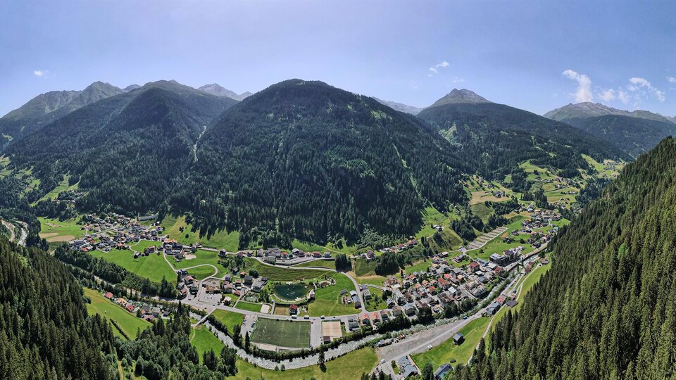 A beautiful panoramic view of a valley surrounded by high mountains. The landscape is green with forests and a small village in the background.
