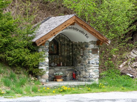 A small stone chapel with a wooden roof stands in nature. Surrounded by trees and grass, it offers a peaceful retreat.