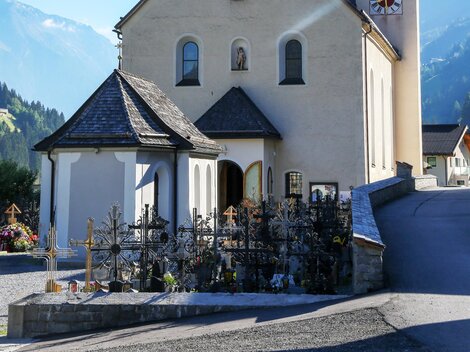 A picturesque church with a distinctive tower in the Alps. In the foreground, there is a small cemetery.