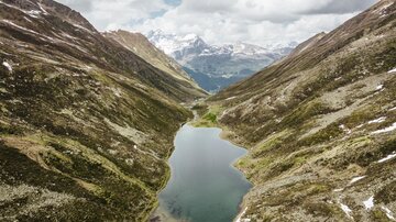 An idyllic mountain landscape with a clear lake in the center. Surrounded by green hills and majestic mountains under a cloudy sky.
