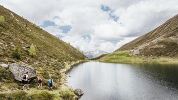 A peaceful landscape with a clear lake, surrounded by mountains. Hikers explore the green surroundings under a cloudy sky.