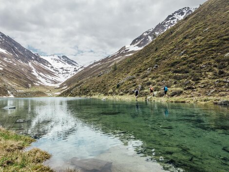 A quiet lake in the mountains, surrounded by snow-capped peaks. Hikers stand by the shore and enjoy nature.