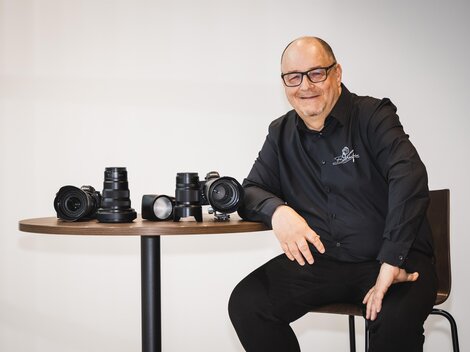 A smiling man sits at a table and presents several cameras and lenses. He is wearing a black shirt and appears friendly and competent. | © Bernhard Gruber – Photography