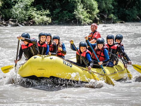 A group of eight people is paddling in a yellow raft on a river. They are wearing helmets and life jackets and having a lot of fun.