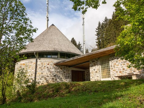 A modern building in a round shape with a pointed roof and window fronts. Surrounded by trees and green areas in a rural setting. | © Bernhard Gruber – Photography