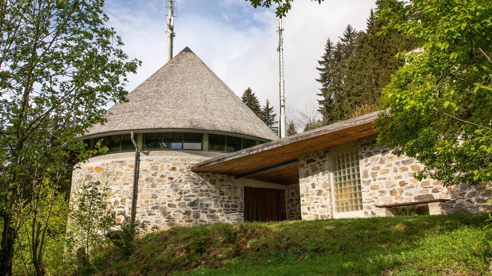 A modern building in a round shape with a pointed roof and window fronts. Surrounded by trees and green areas in a rural setting. | © Bernhard Gruber – Photography
